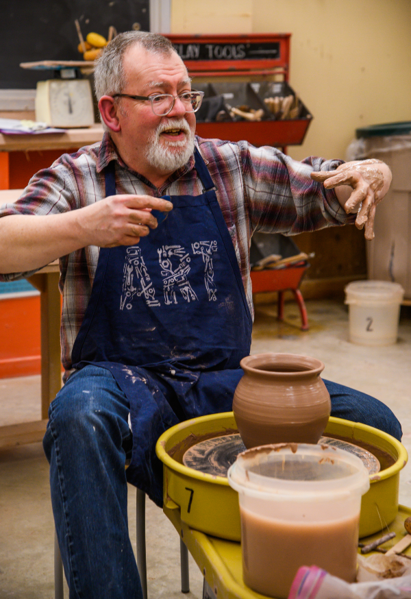 Instructor Greg Lamont wearing apron sitting at a pottery wheel with pot in front of him. Hands are lifted as if demonstrating a skill